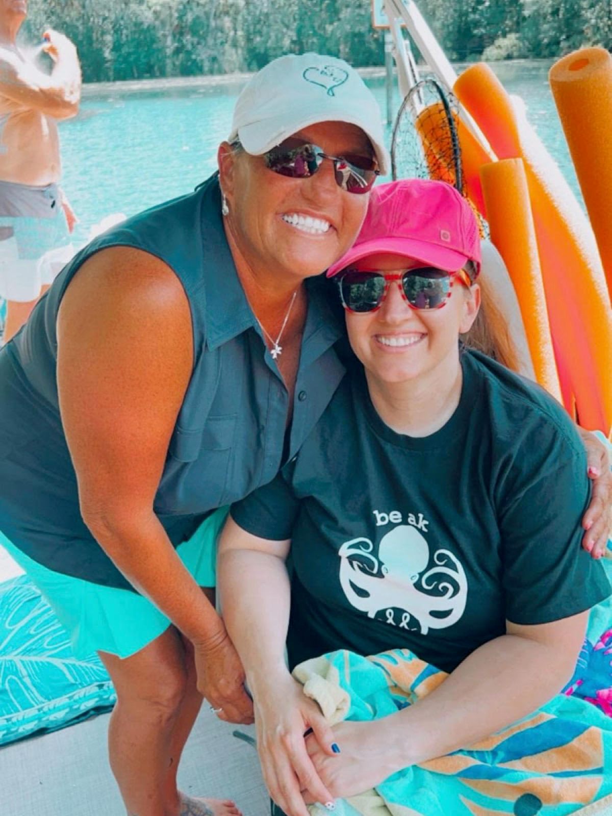 Two women smiling on a boat with pool noodles and water background.