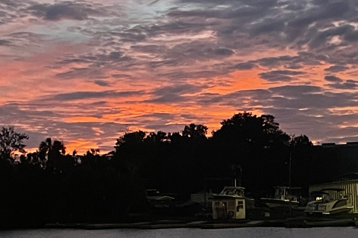 Sunset over water with boats and silhouetted trees, pink and orange sky with clouds.