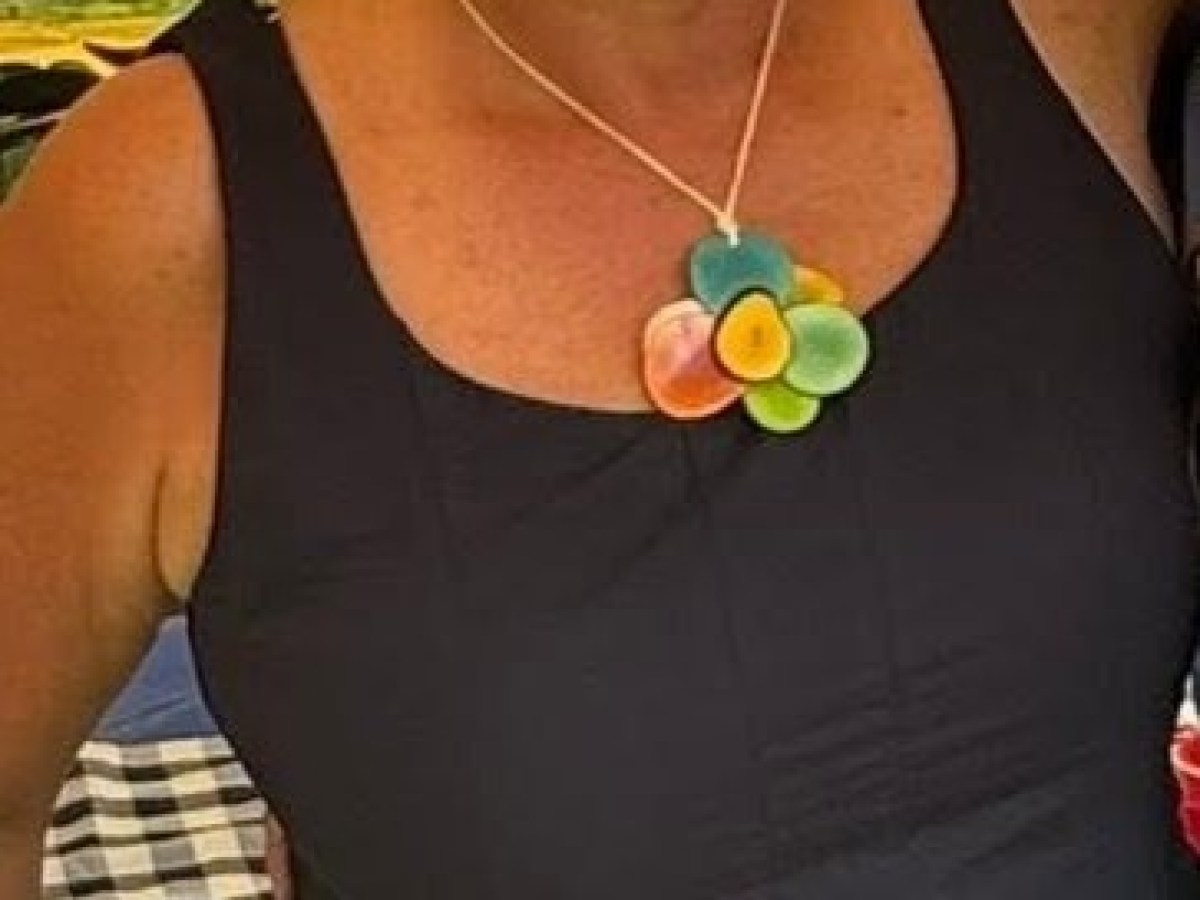 Smiling person in sunglasses and a black dress with colorful necklace outdoors.
